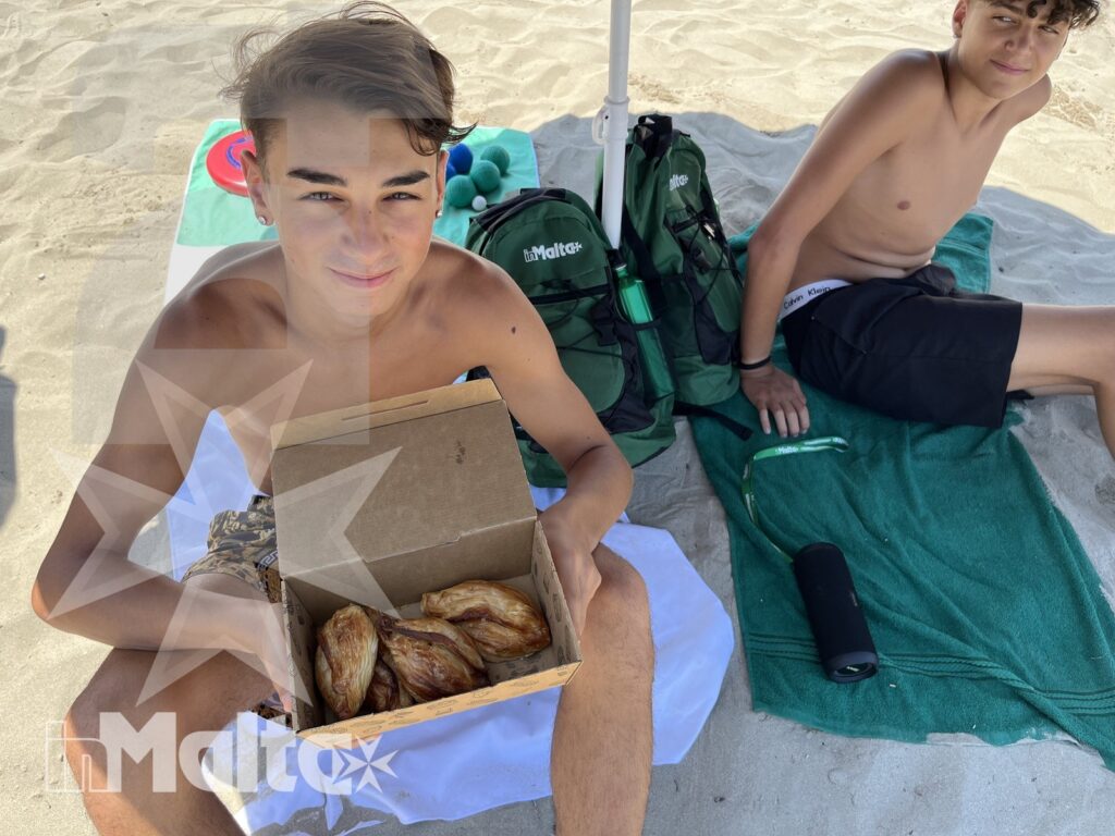 Due studenti seduti sulla spiaggia mostrano una scatola di prodotti da forno sotto l'ombrellone, in un momento di condivisione.