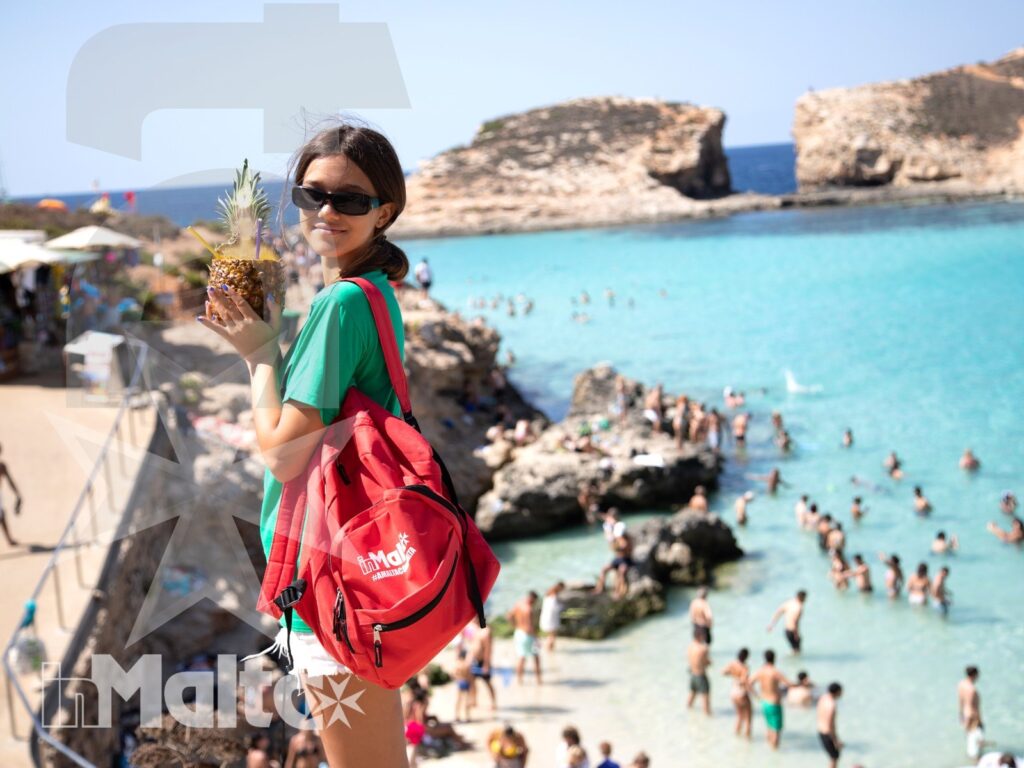 Studentessa inMalta davanti alla baia durante l'escursione Studentessa con zaino rosso e ananas osserva la baia turchese affollata durante un'escursione sulla costa di Malta.