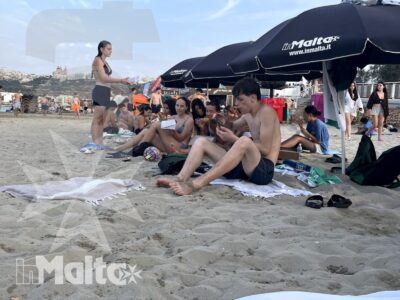 Gruppo numeroso di studenti si rilassa sotto gli ombrelloni sulla spiaggia di Mellieħa Bay, con il panorama urbano alle spalle.
