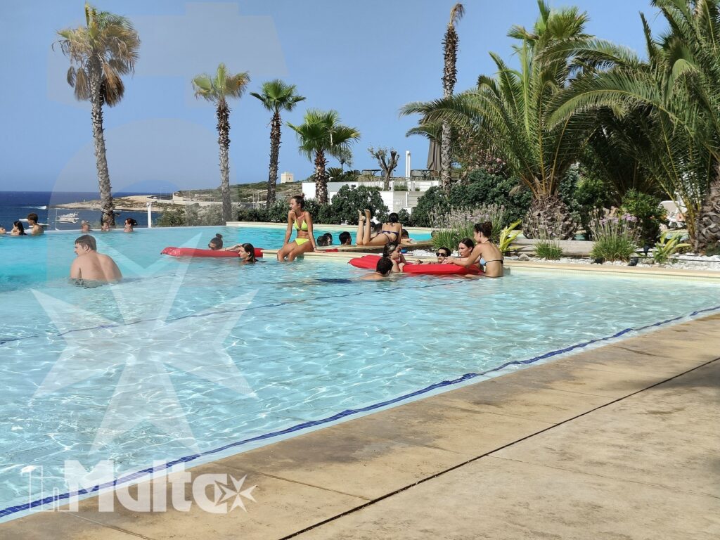 Giovani bagnanti si divertono su materassini colorati nella piscina all'aperto, tra palme e mare durante una vacanza studio.