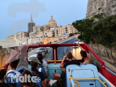 Passeggeri sull'autobus scoperto ammirano La Valletta, con bastioni storici, cupola e campanili visibili nel panorama.