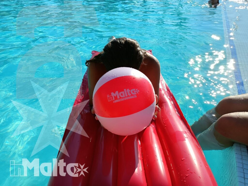 Persona su un materassino rosso in piscina, con pallone dedicato a Malta davanti al volto e acqua turchese intorno.