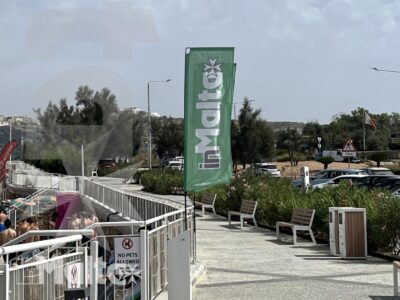 Percorso pedonale recintato conduce all'ingresso di una struttura a Malta, con area ricreativa e ombrelloni sullo sfondo.