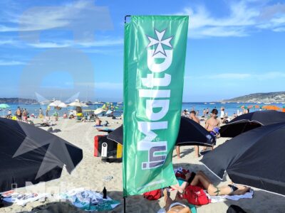 Spiaggia sabbiosa affollata con bagnanti, ombrelloni e una bandiera verde in primo piano, nell'atmosfera estiva di Mellieħa Bay.