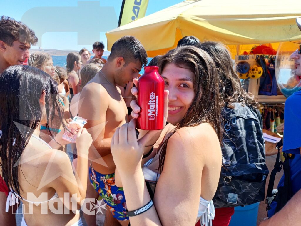 Partecipanti inMalta durante una giornata al mare a Gozo Studentessa sorridente mostra una borraccia sulla spiaggia, circondata da partecipanti e da un chiosco durante un'attività inMalta.