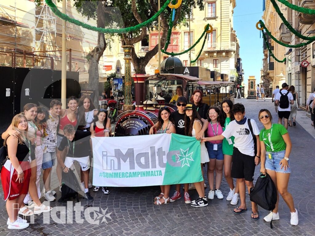 Gruppo internazionale di studenti posa con uno striscione in una strada storica decorata di Malta durante un'escursione urbana.