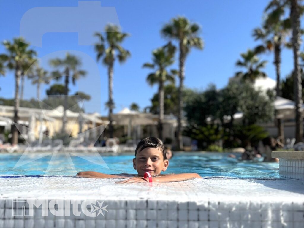 Bambino sorridente si appoggia al bordo della piscina, con acqua turchese e palme sfocate sullo sfondo.