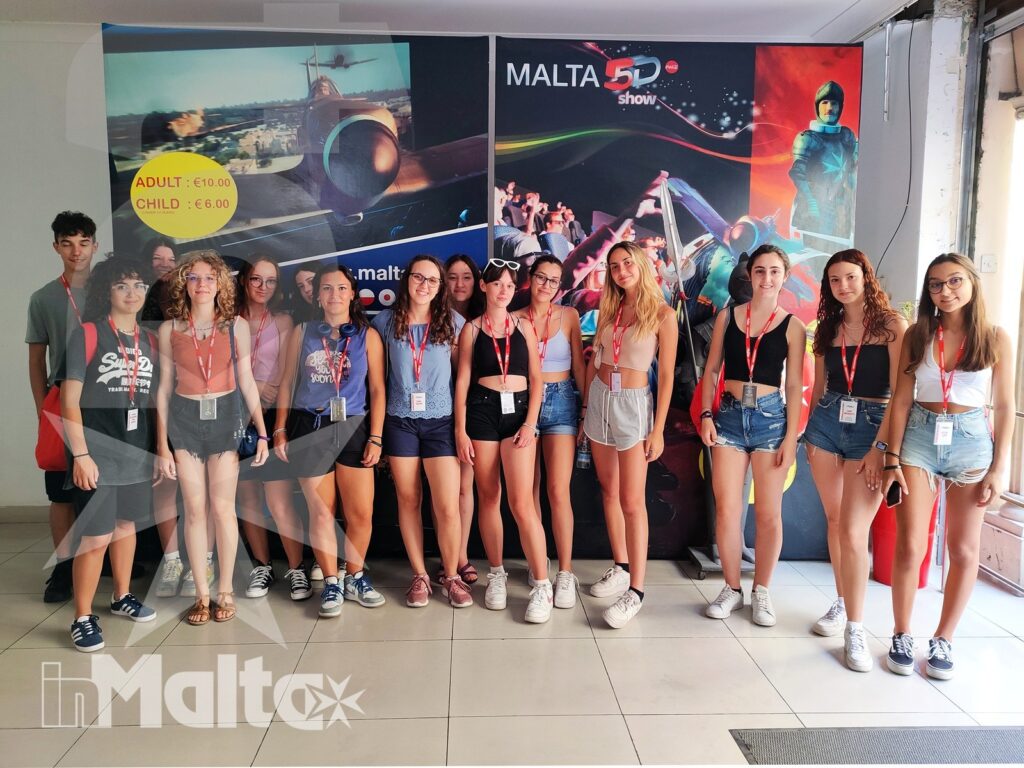 Gruppo internazionale di studenti posa in ambiente interno davanti a pannelli promozionali dedicati a Malta, con badge da partecipanti.