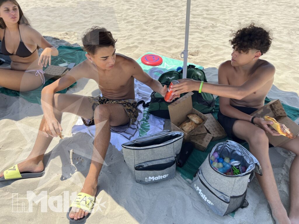 Tre giovani condividono snack e bevande sotto un ombrellone, durante una pausa conviviale in spiaggia per un'esperienza inMalta.