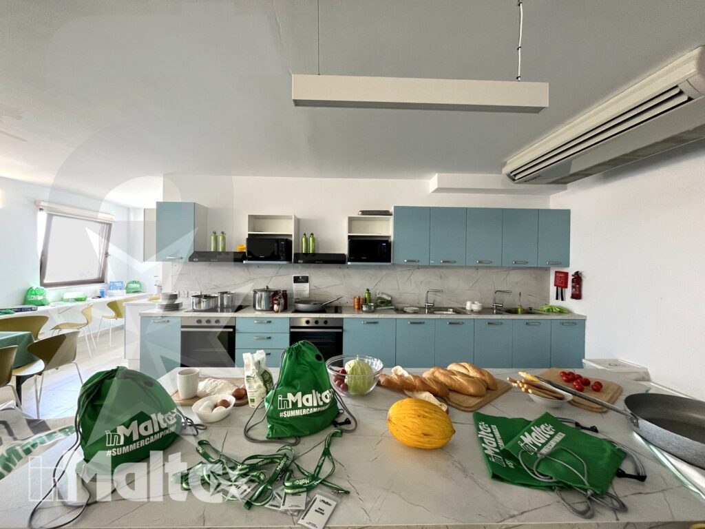 Cucina comune moderna con bancone centrale, pane, frutta e borse verdi con marchio Malta, pronta per preparare i pasti.