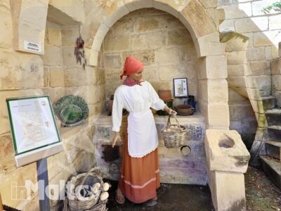 Donna in costume storico attinge acqua da una fontana medievale in pietra durante una rievocazione culturale a Malta.