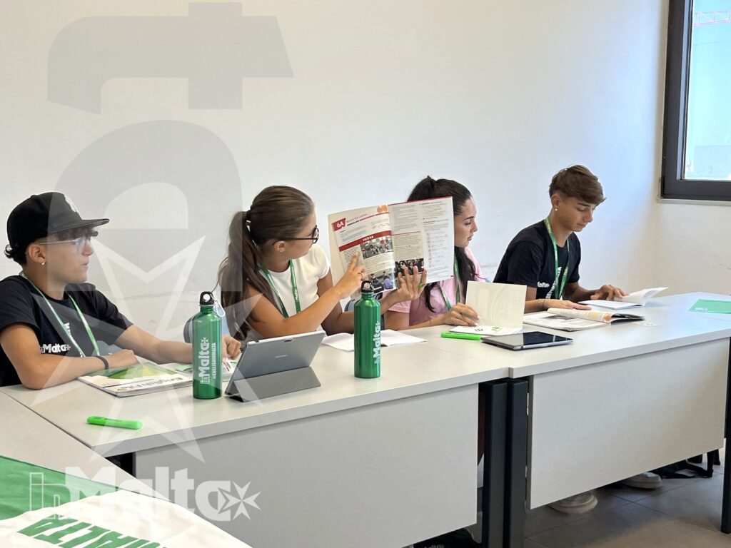 Studenti leggono dispense e lavorano insieme con tablet e materiali cartacei durante una sessione formativa organizzata inMalta.