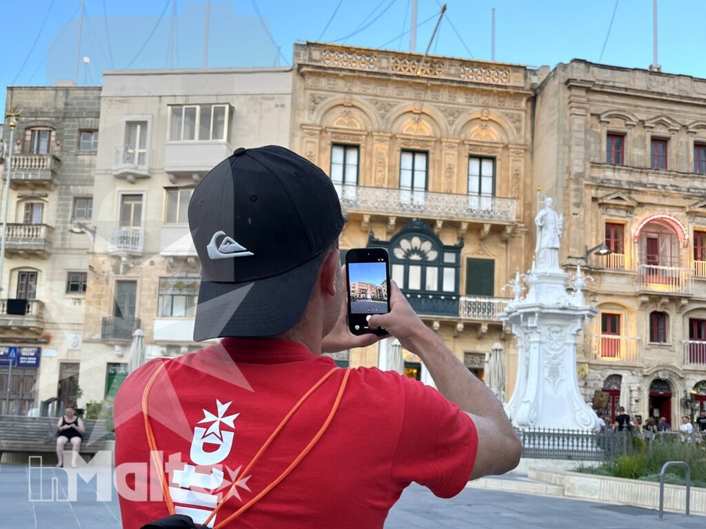 Studente inMalta fotografa una piazza durante il tour culturale Studente fotografa con lo smartphone una fontana monumentale e gli edifici storici di una piazza maltese.