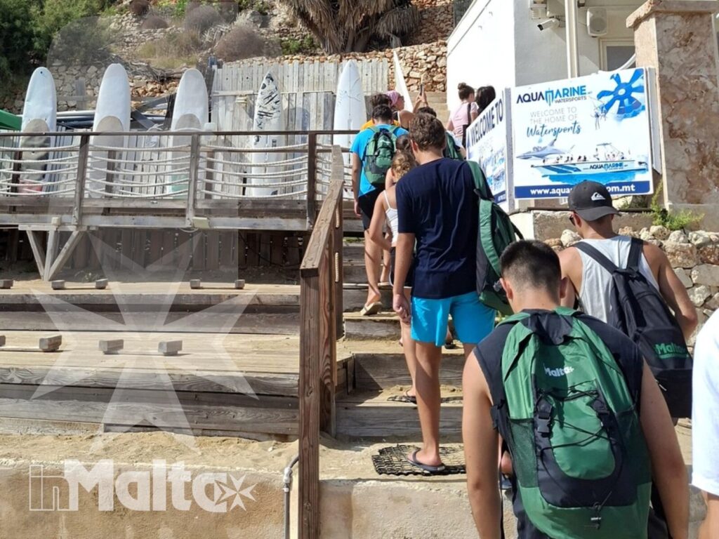 Gruppo di studenti con zaini sale una scala verso una struttura balneare, tra palme, hotel e paesaggio mediterraneo sulla costa maltese.