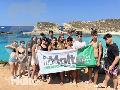 Gruppo internazionale di studenti posa con lo striscione del programma davanti al mare turchese e alla costa rocciosa maltese.