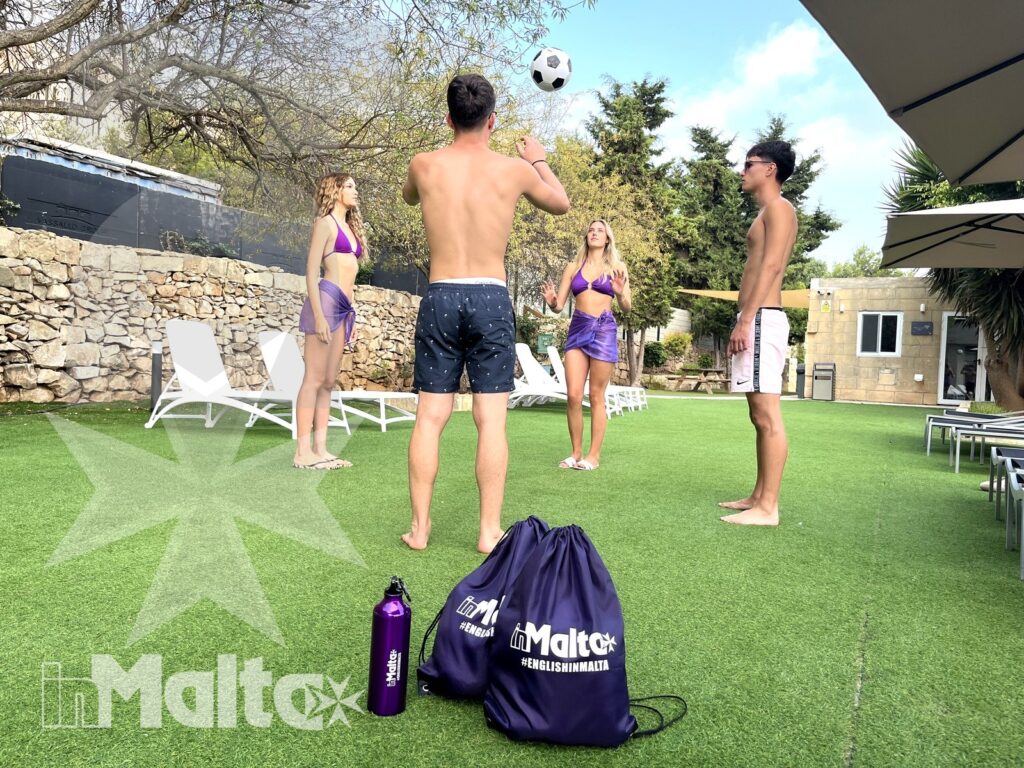 Attività all'aperto per studenti durante il soggiorno inMalta Quattro studenti internazionali giocano con un pallone sul prato della struttura, tra abbigliamento estivo, borse e attrezzatura del tour operator.