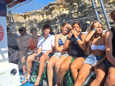 Gli studenti condividono una gita in barca lungo la costa rocciosa di Malta, ripresi insieme sotto il tendalino rosso.