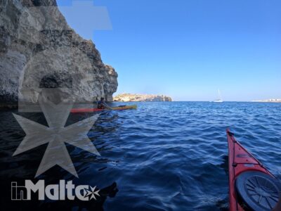 Un kayak rosso attraversa il mare limpido davanti a una scogliera con grotta, mentre una barca a vela appare sullo sfondo.
