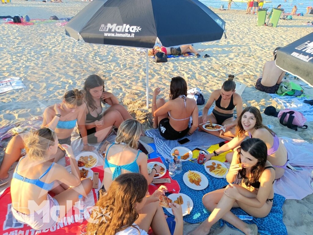 Studentesse in vacanza studio pranzano sulla spiaggia Gruppo di studentesse condivide pranzo e bevande sulla spiaggia, seduta su una stuoia sotto l'ombrellone durante una vacanza studio a Malta.