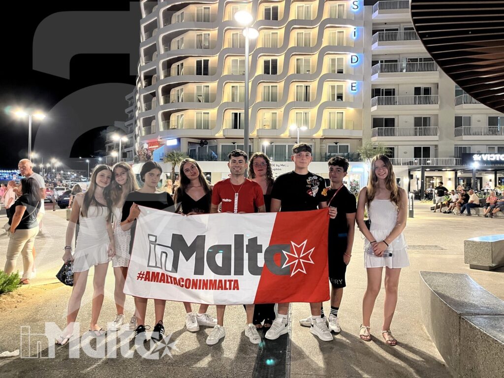 Studenti sorridenti posano con la bandiera di Malta in una vivace piazza notturna, tra edifici moderni e luci urbane.
