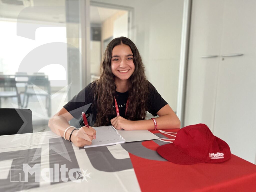 Studentessa sorridente scrive su un quaderno a un tavolo, con cappellino rosso e materiali promozionali dell'attività formativa.