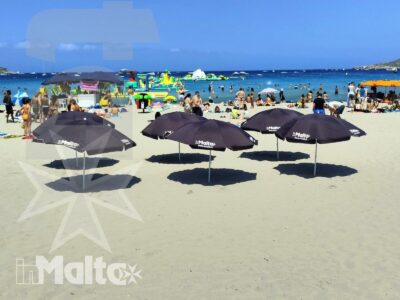 Tre ombrelloni neri in primo piano incorniciano la spiaggia affollata e il mare blu di Mellieħa Bay.