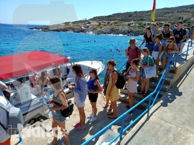 Gruppo numeroso di studenti sale su una barca dal pontile, accanto a un litorale roccioso con bandiera maltese.