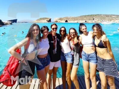 Sette studentesse sorridono sul pontile, davanti al mare turchese e alla costa rocciosa maltese durante un'uscita di gruppo.