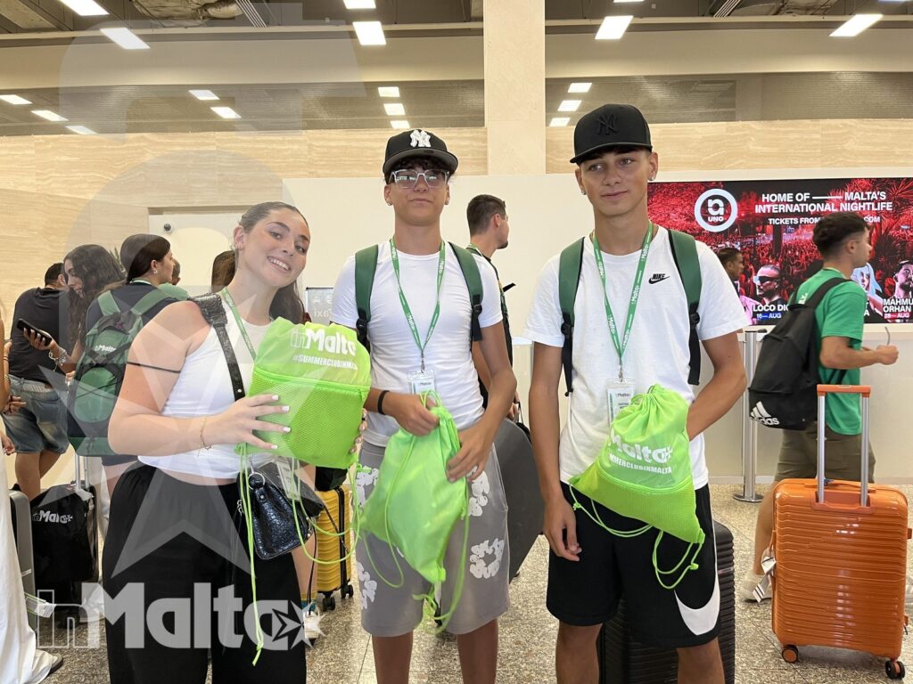 Studenti inMalta all'arrivo con borse e valigie Tre studenti sorridenti posano nell'area arrivi di Malta con borse verdi del tour e una valigia arancione.