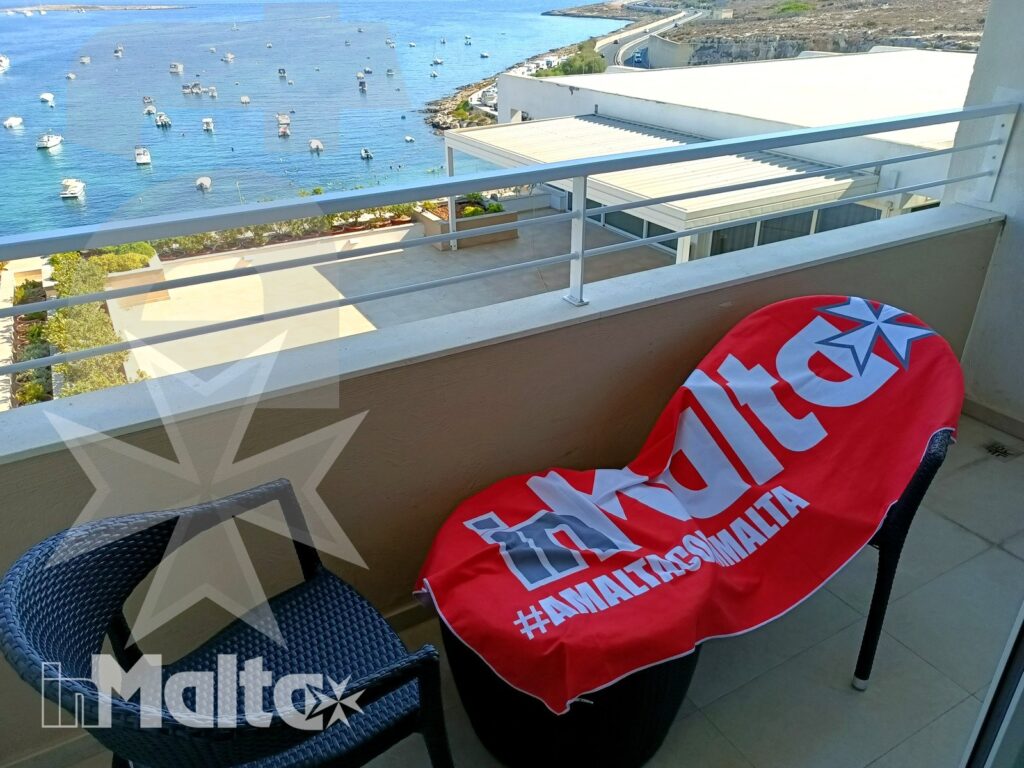 Balcone arredato con asciugamano rosso brandizzato in primo piano e vista sulla baia di Malta con barche