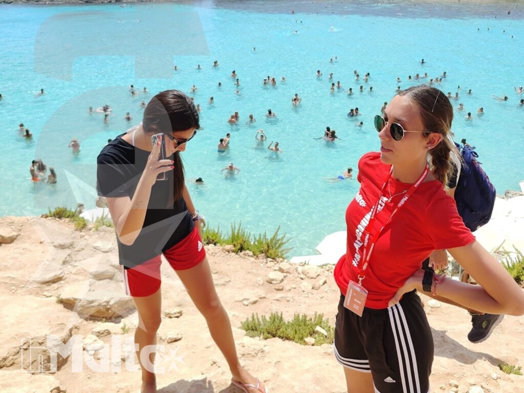 Due studentesse in escursione alla laguna di Comino Due studentesse posano davanti alla laguna turchese affollata e alla costa rocciosa durante un'escursione a Comino.