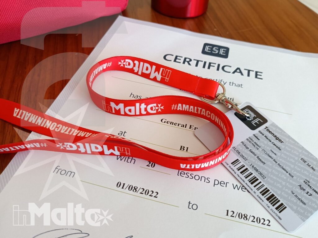 Certificato di frequenza, tessera identificativa e laccetto con riferimenti a Malta disposti su un tavolo di legno.