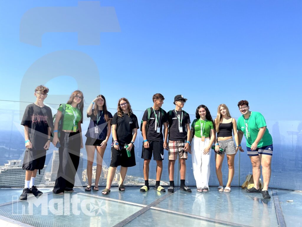 Gruppo multiculturale di giovani posa su una terrazza riflettente con parapetto trasparente e skyline urbano, durante un viaggio organizzato.