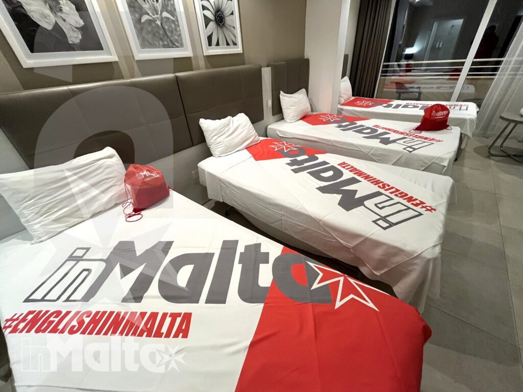 Letti singoli allineati con bandiere maltesi, grafiche promozionali e decorazioni rosse in una camera condivisa per giovani ospiti.