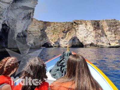 Studenti con giubbotti salvagente osservano grotte e scogliere calcaree da una barca durante un'escursione costiera a Malta.