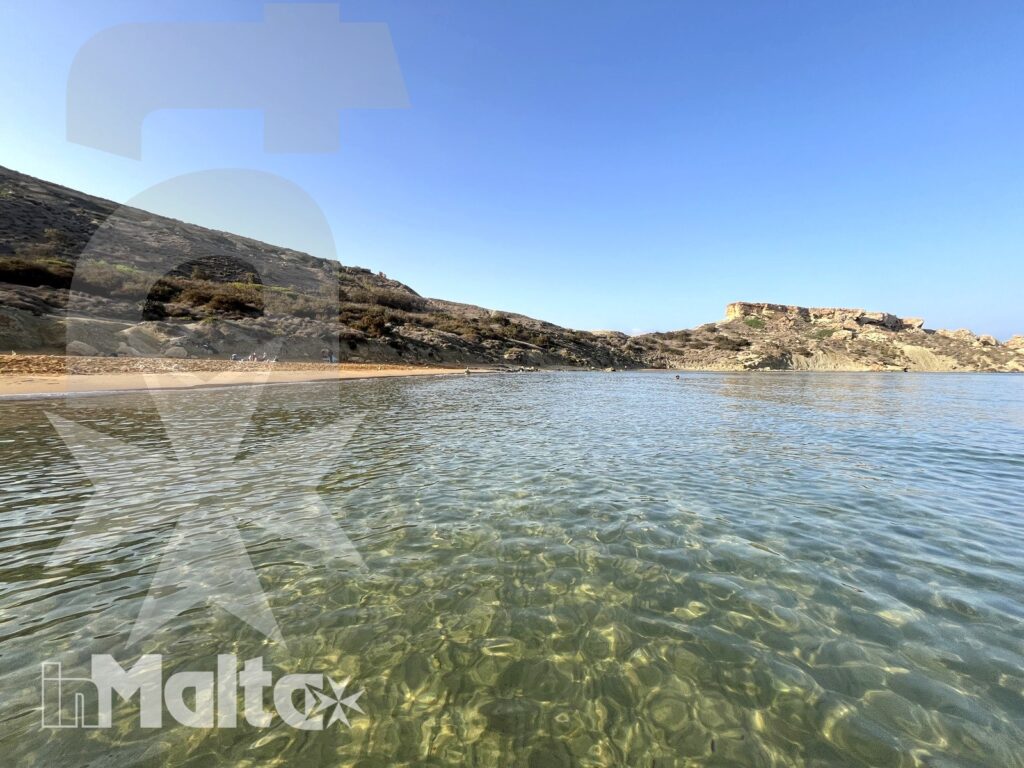 Acqua marina trasparente lascia vedere il fondale roccioso di una baia maltese, incorniciata da costa sabbiosa e scogliere basse.