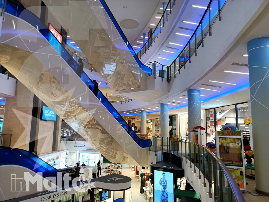 Atrio luminoso di un moderno centro commerciale multipiano, con scale mobili blu, negozi e ampie vetrate.