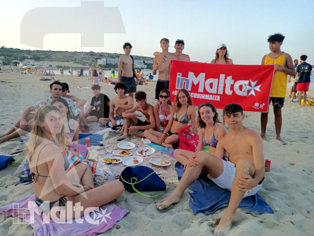 Studenti inMalta durante un picnic sulla spiaggia Gruppo internazionale di studenti prende parte a un picnic sulla sabbia, con la bandiera di Malta e la spiaggia sullo sfondo.