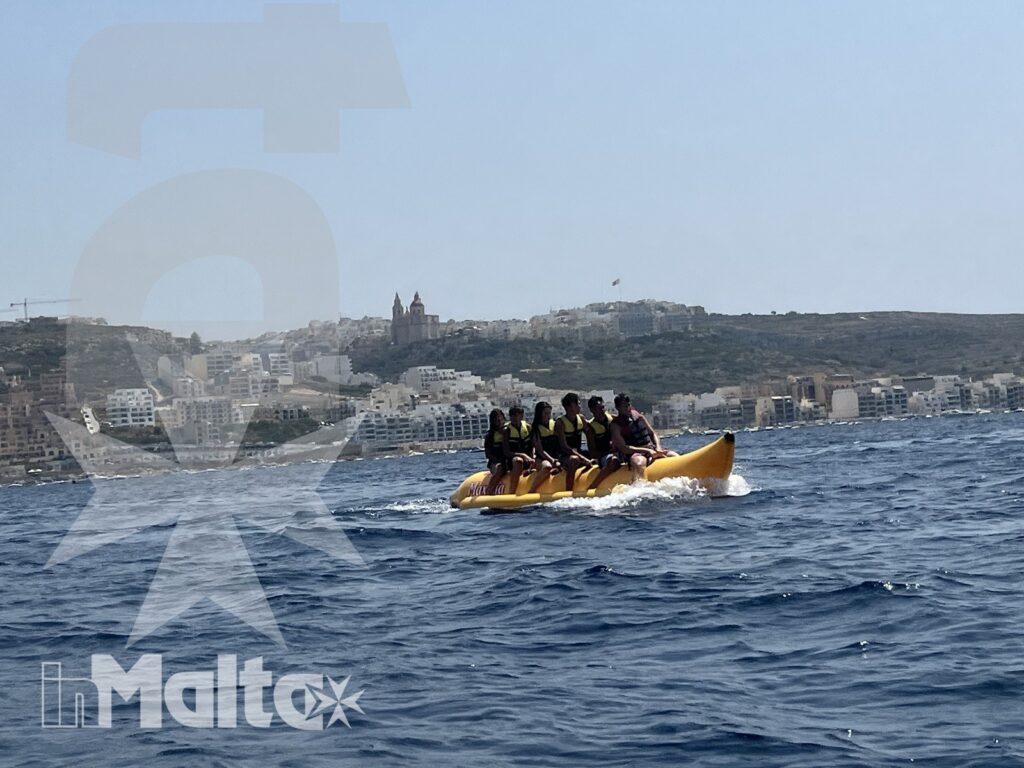 Gruppo di giovani naviga su un banana boat giallo davanti a una città costiera, tra mare mosso e spruzzi.