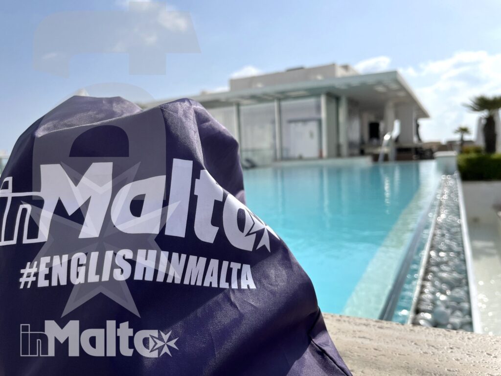 Borsa English in Malta a bordo piscina Borsa viola brandizzata in primo piano davanti alla piscina turchese e alla struttura moderna, simbolo dell'esperienza English in Malta.