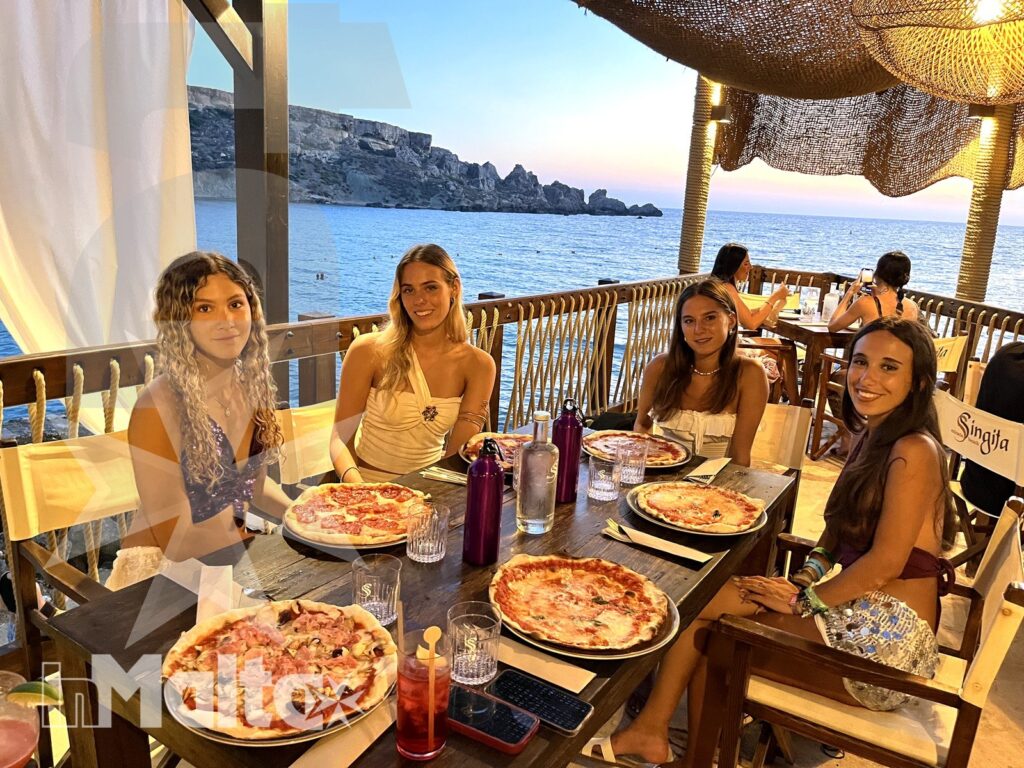 Un gruppo di studentesse sorride a tavola durante una cena in pizzeria, con la costa maltese sullo sfondo.