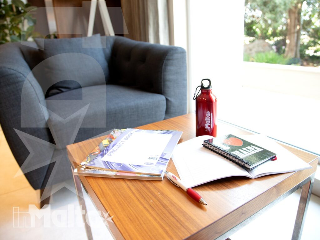 Poltrona accanto a un tavolino con brochure, quaderno, penna e borraccia, in un angolo studio affacciato sul verde.