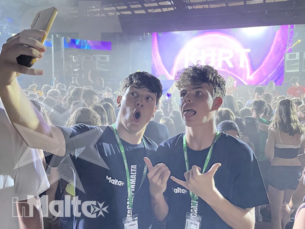 Due studenti sorridenti scattano un selfie davanti a un palco illuminato, con la folla alle spalle durante una serata musicale.