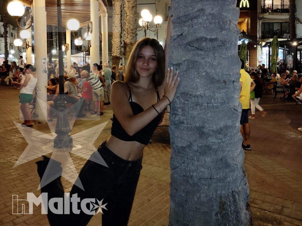 Studentessa accanto a una palma in una piazza serale affollata, tra locali, ristoranti e luci decorative durante la passeggiata a Bugibba.