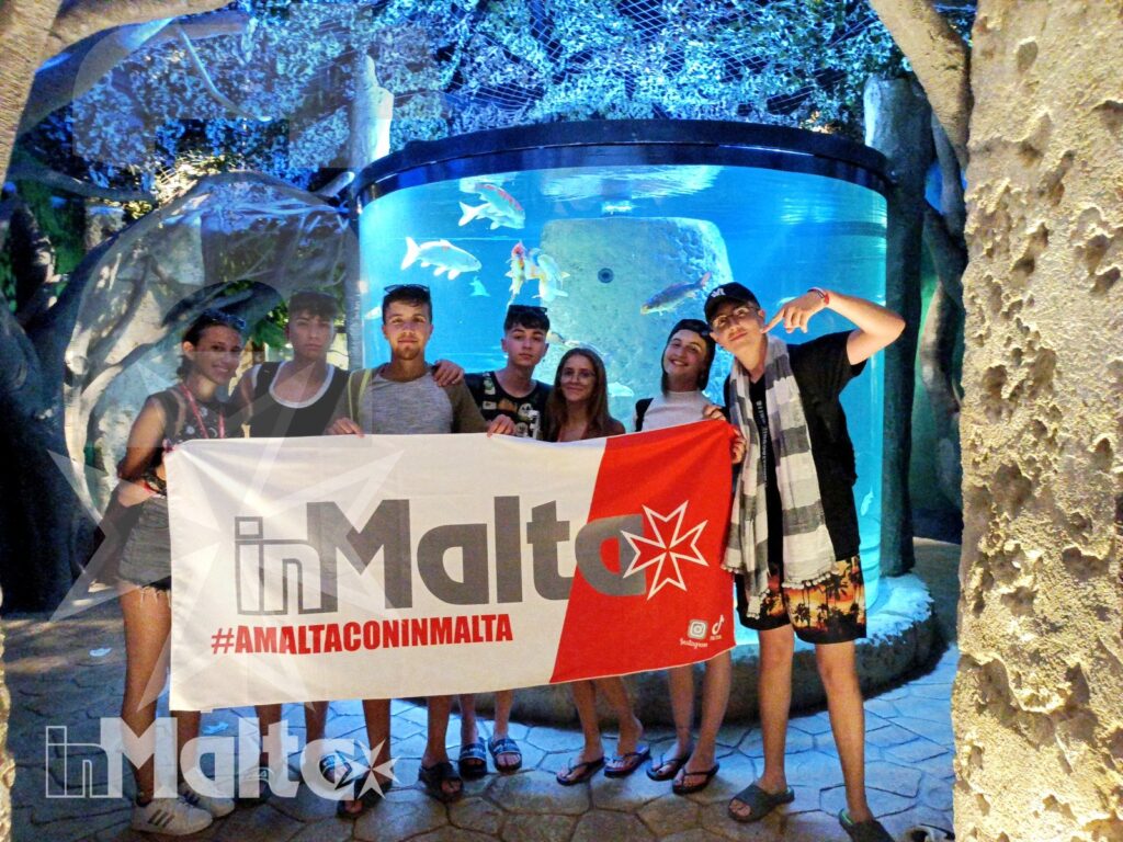 Studenti inMalta in posa davanti alle vasche dell'acquario Gruppo di studenti posa con uno striscione inMalta davanti a una vasca illuminata durante la visita all'acquario.