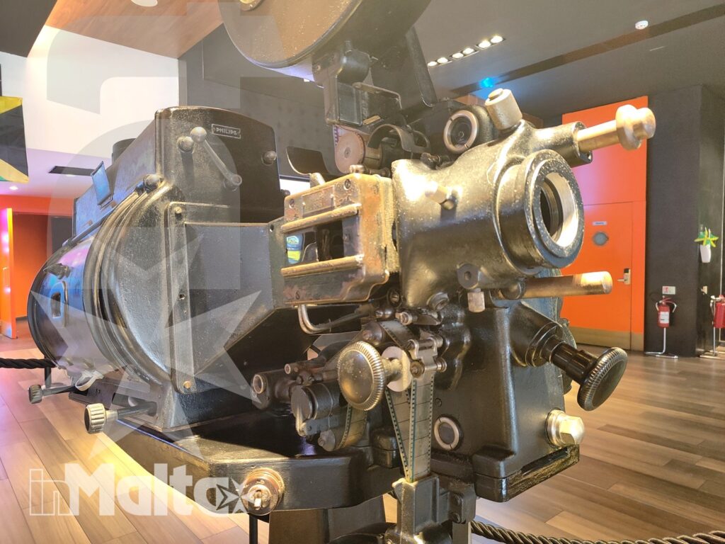 Dettaglio di un proiettore cinematografico storico esposto in una sala moderna con illuminazione colorata e pavimento in legno.