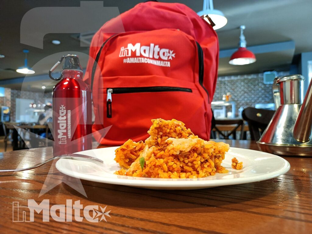 Piatto di riso speziato su un tavolo con zaino e borracce brandizzate, in una caffetteria frequentata durante il soggiorno a Malta.