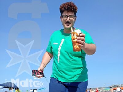 Uno studente cammina sulla sabbia con una bevanda in mano, tra bagnanti e mare nella vivace spiaggia di Mellieħa Bay.