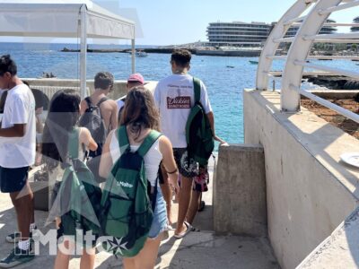 Studenti con zaini percorrono la passerella verso il pontile, tra mare limpido, costa rocciosa e strutture sulla riva.