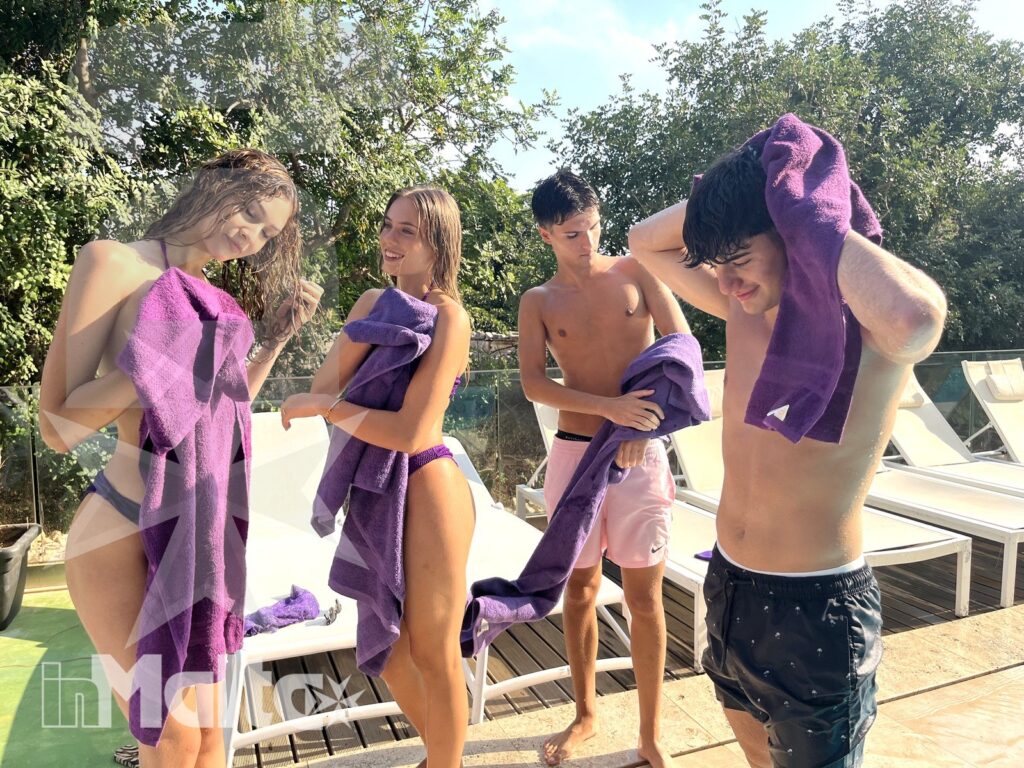 Studenti inMalta si rilassano a bordo piscina dopo il bagno Quattro studenti sorridono e si asciugano a bordo piscina con asciugamani viola, tra lettini e vegetazione mediterranea durante il soggiorno studio a Malta.
